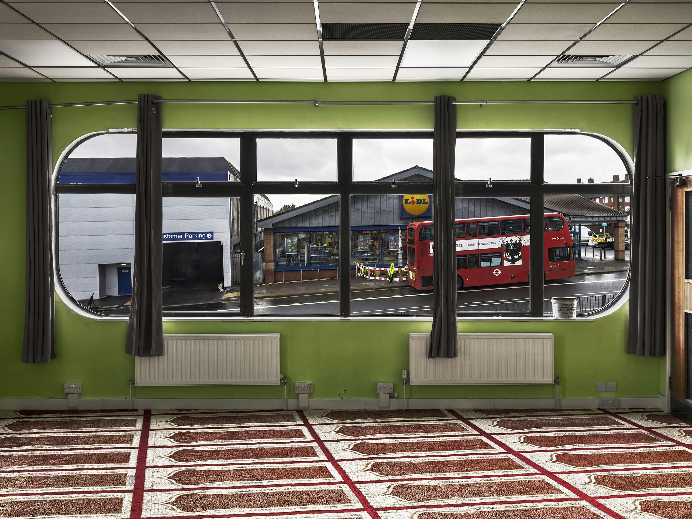 A photograph by Marwan Bassiouni from his 'New British VIews' series taken from the inside of a British Mosque, looking through the window we can see a red bus passing by a Lidl store. 