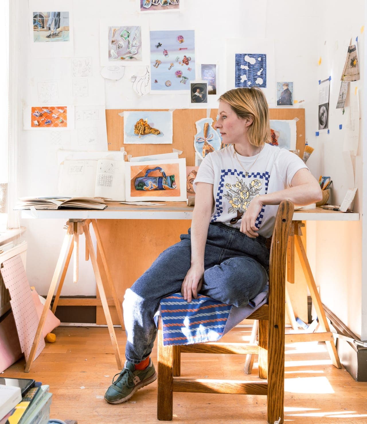 A portrait of Sarah Poots sat in her studio in front of drawings and paintings