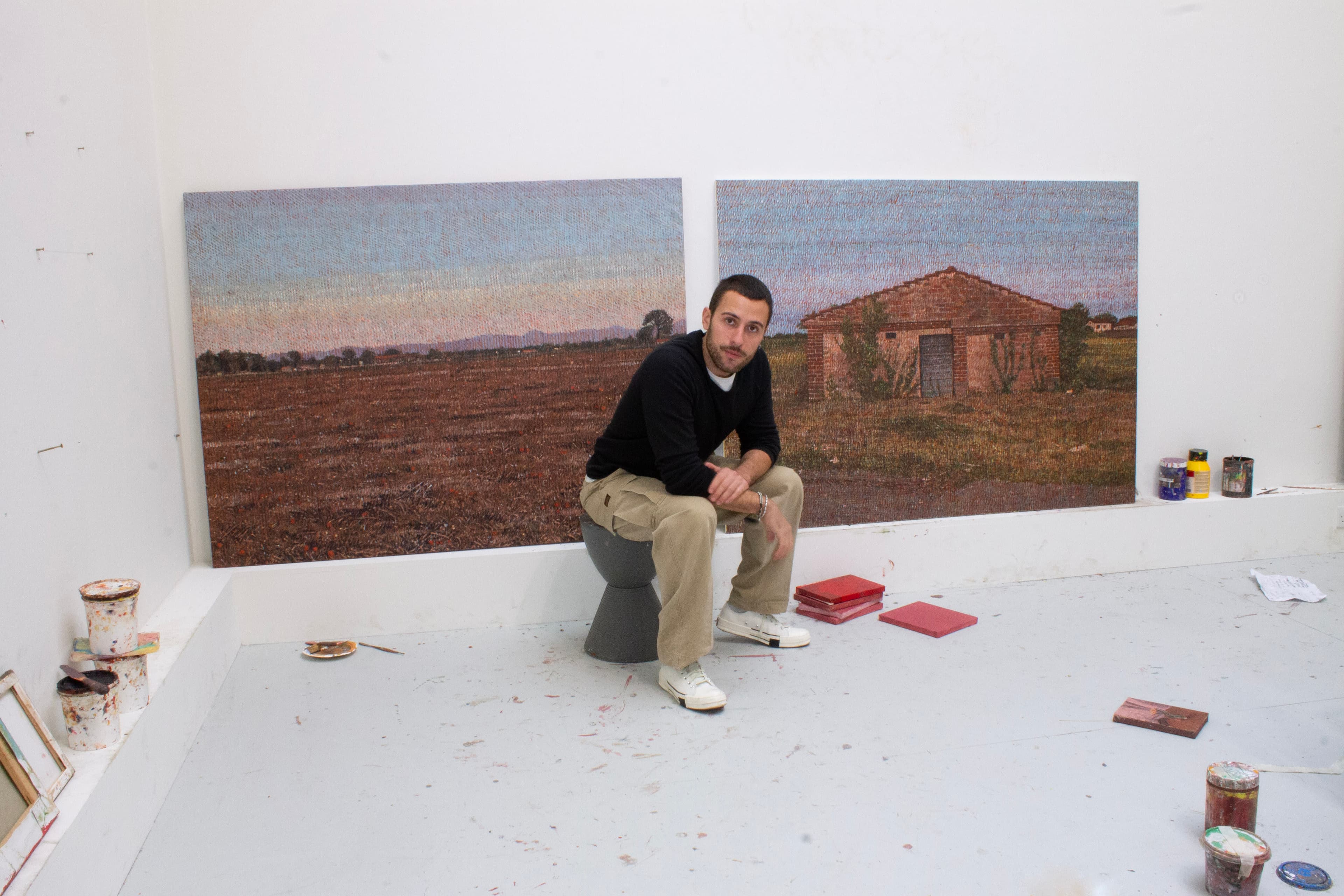 A portrait of Norberto Spina sat in front of two paintings of a landscape and a single storey building