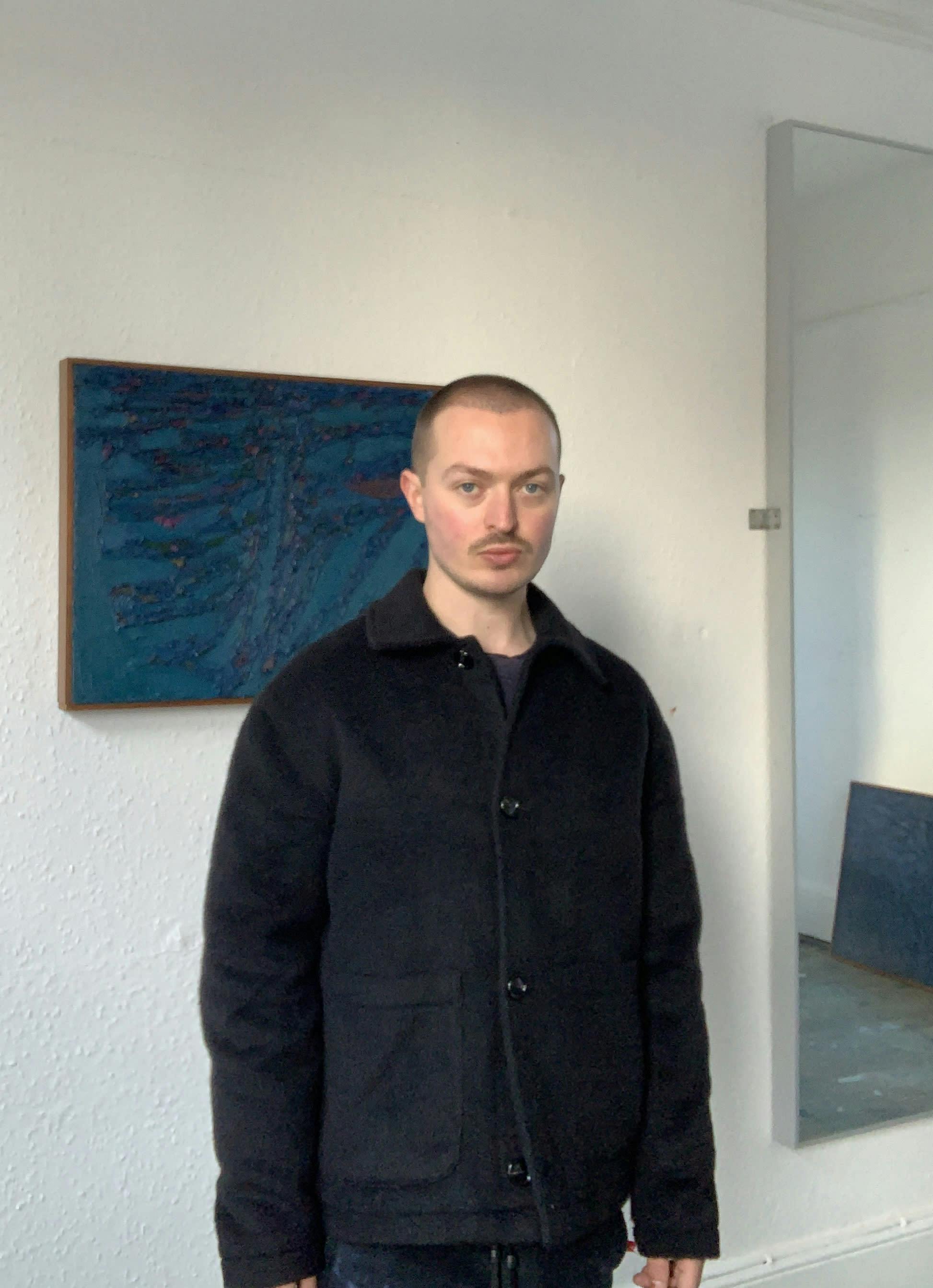 A white man with short brown hair standing in front of a dark blue painting in a white room