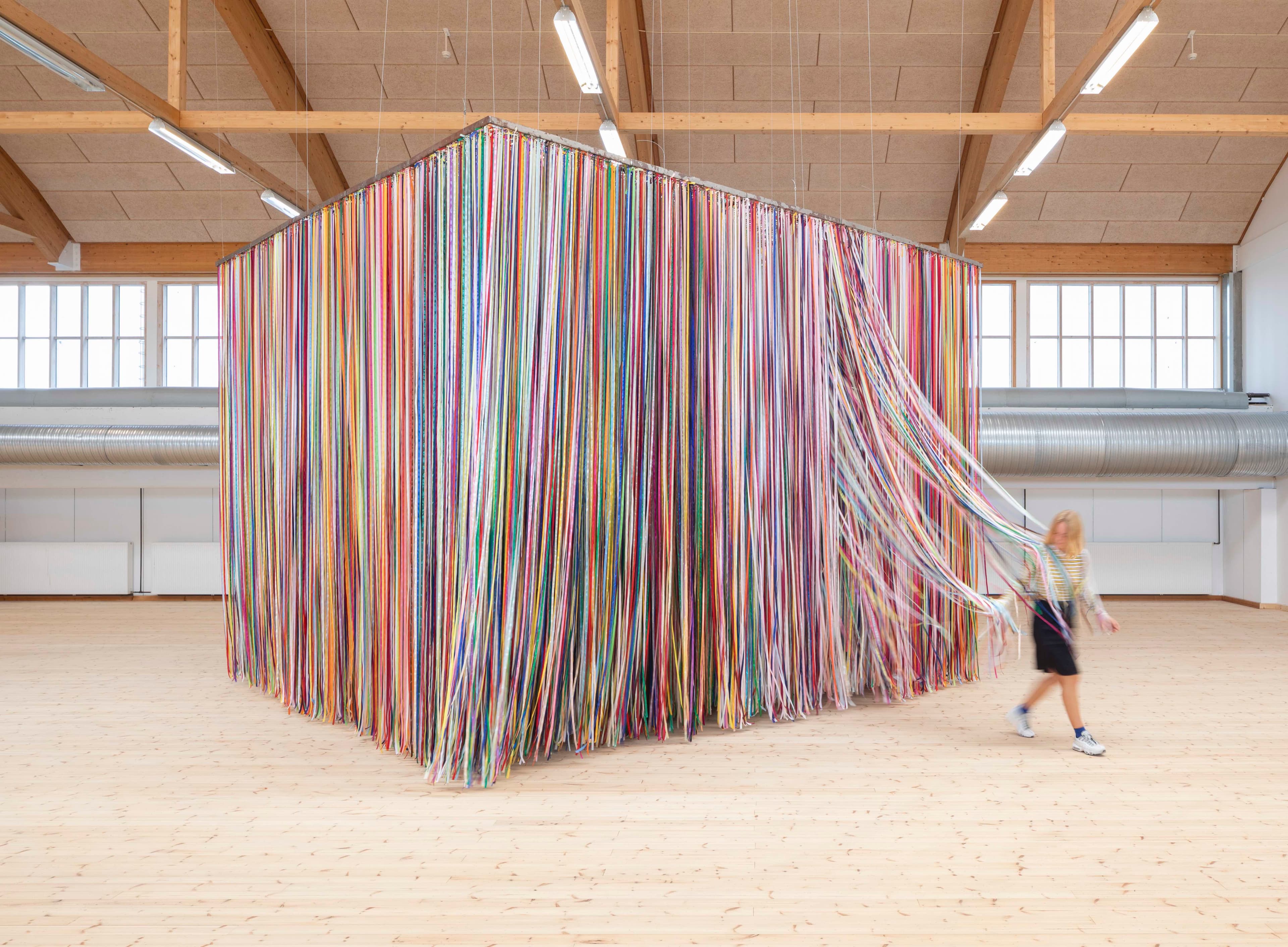 An installation by Jacob Dahlgren comprised of thousands of strands of hanging multi-coloured ribbon creating a giant cube. A young girl is emerging from the work with ribbons trailing behind her