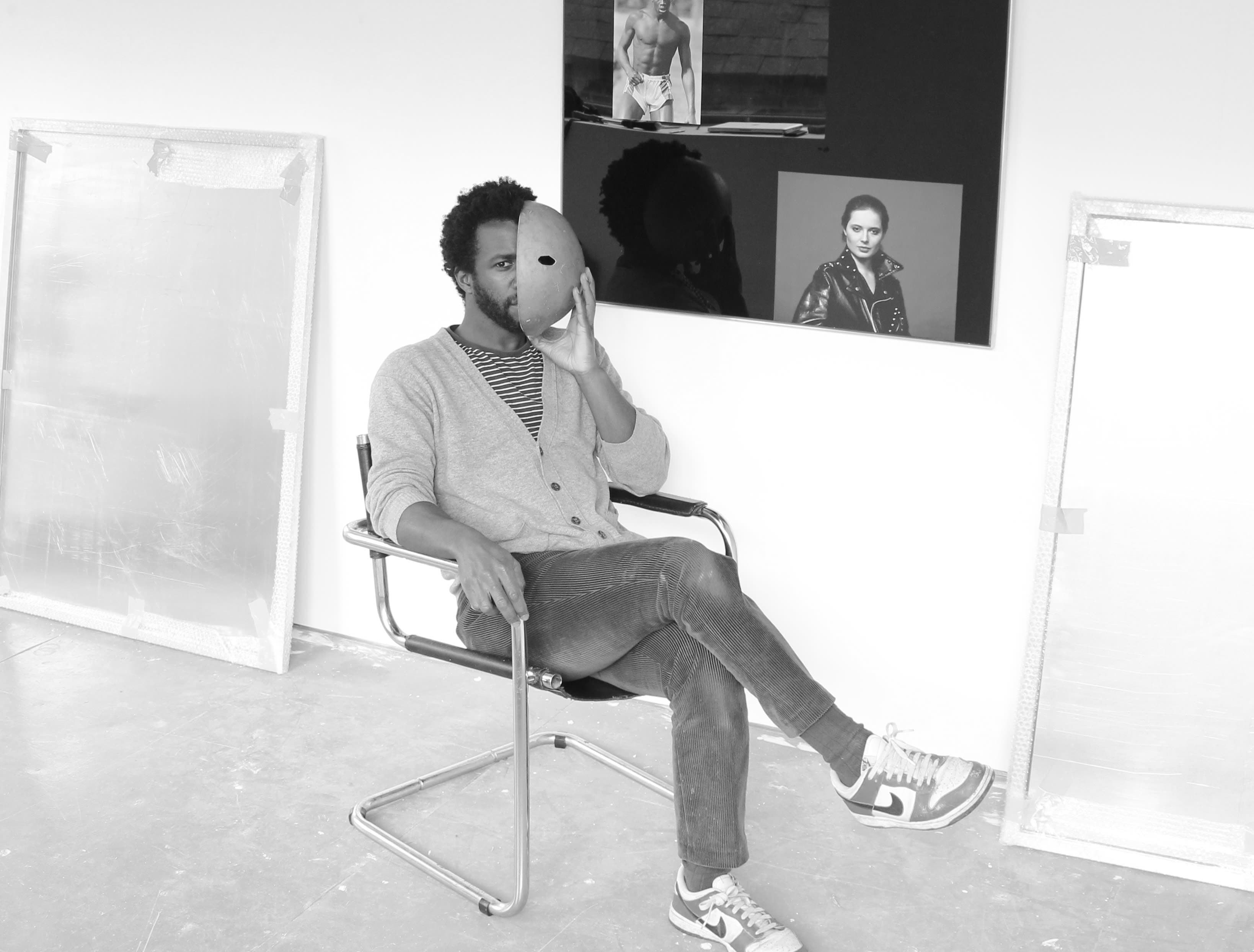 A black and white portrait of Simeon Barclay sat on a chair with his legs crossed and holding a mask over half his face