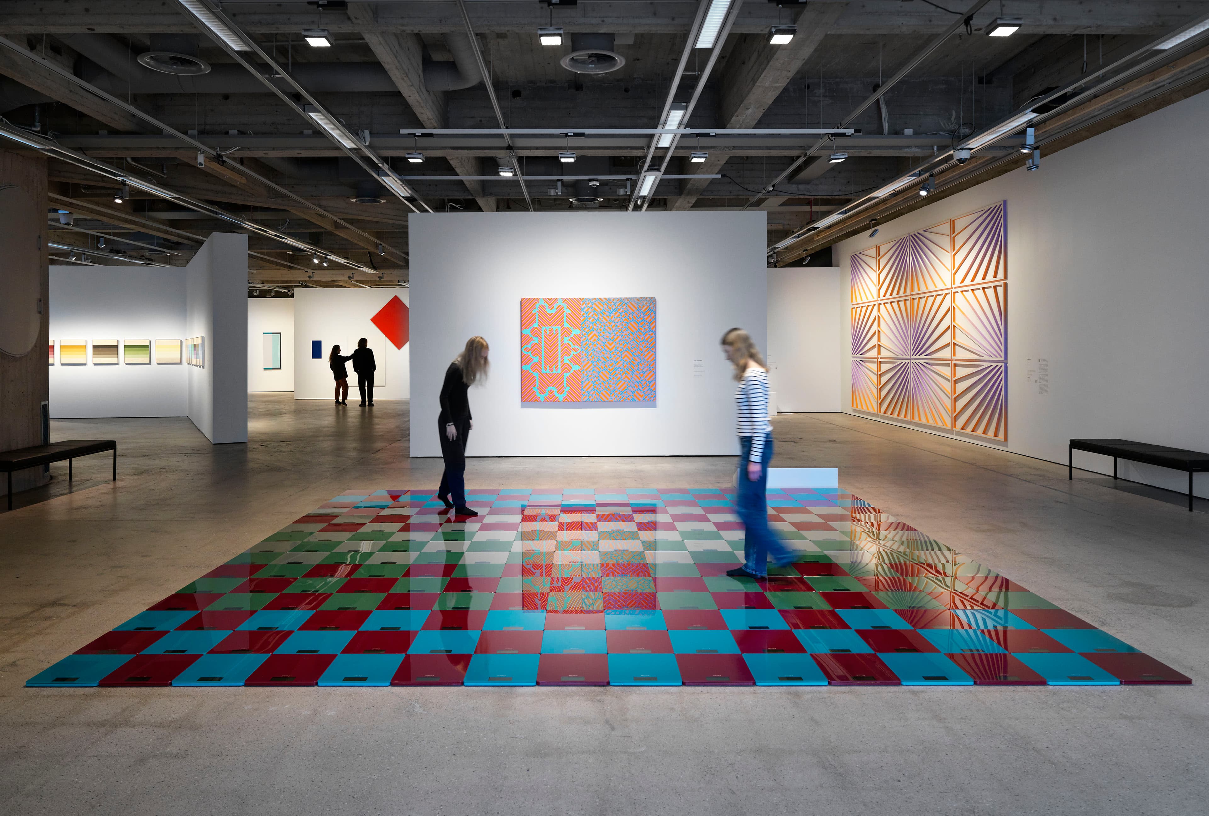 An installation by Jacob Dahlgren featuring hundreds of blue, red and green ikea weighing scales arranged into a square on the floor. Two women walk on top of the artwork.