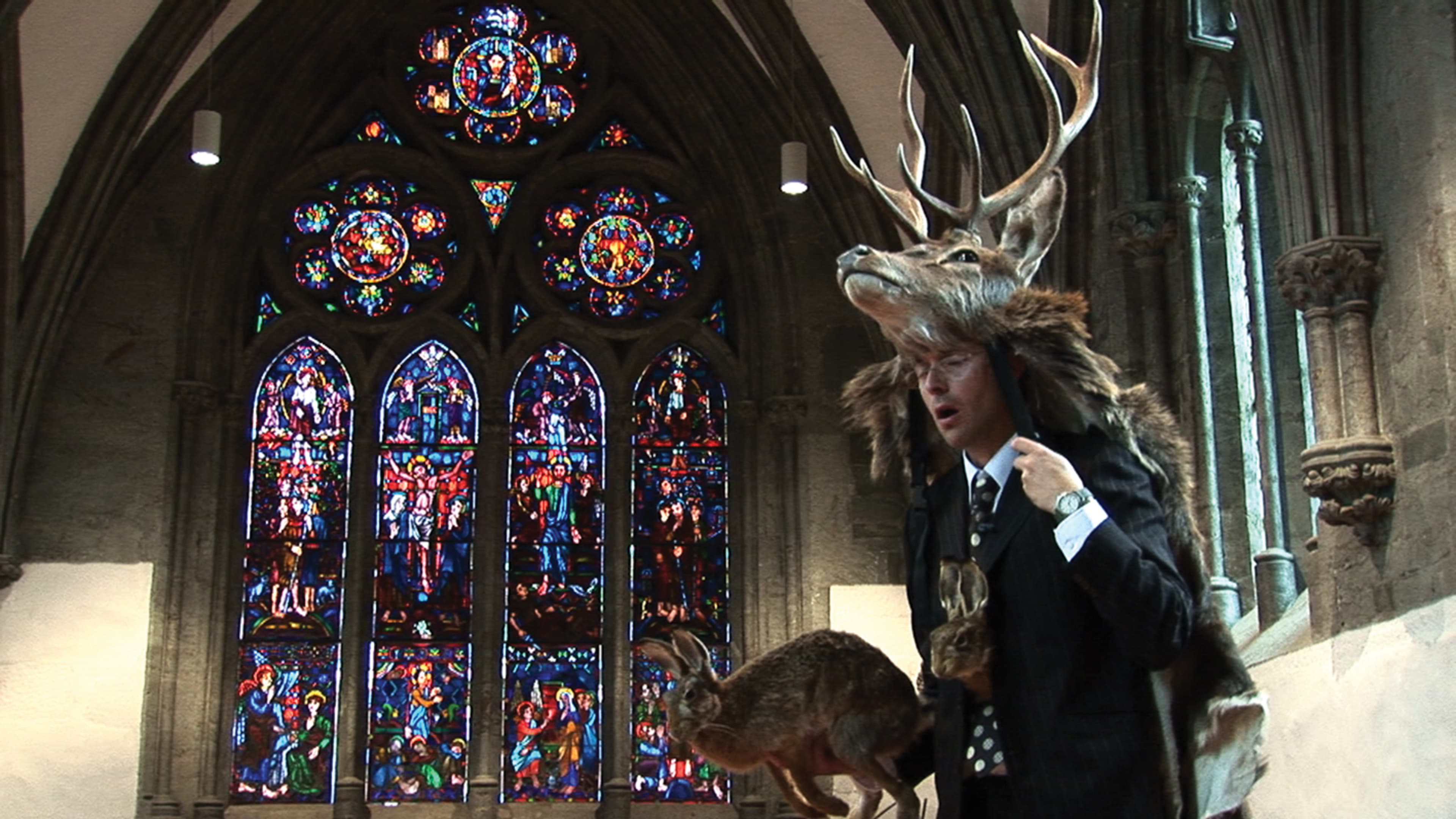 A still from Marcus Coates film 'Radio Shaman' a bespectacled man in a suit holds a stuffed hare and wears a stag's head in front of a stained glass cathedral window. 