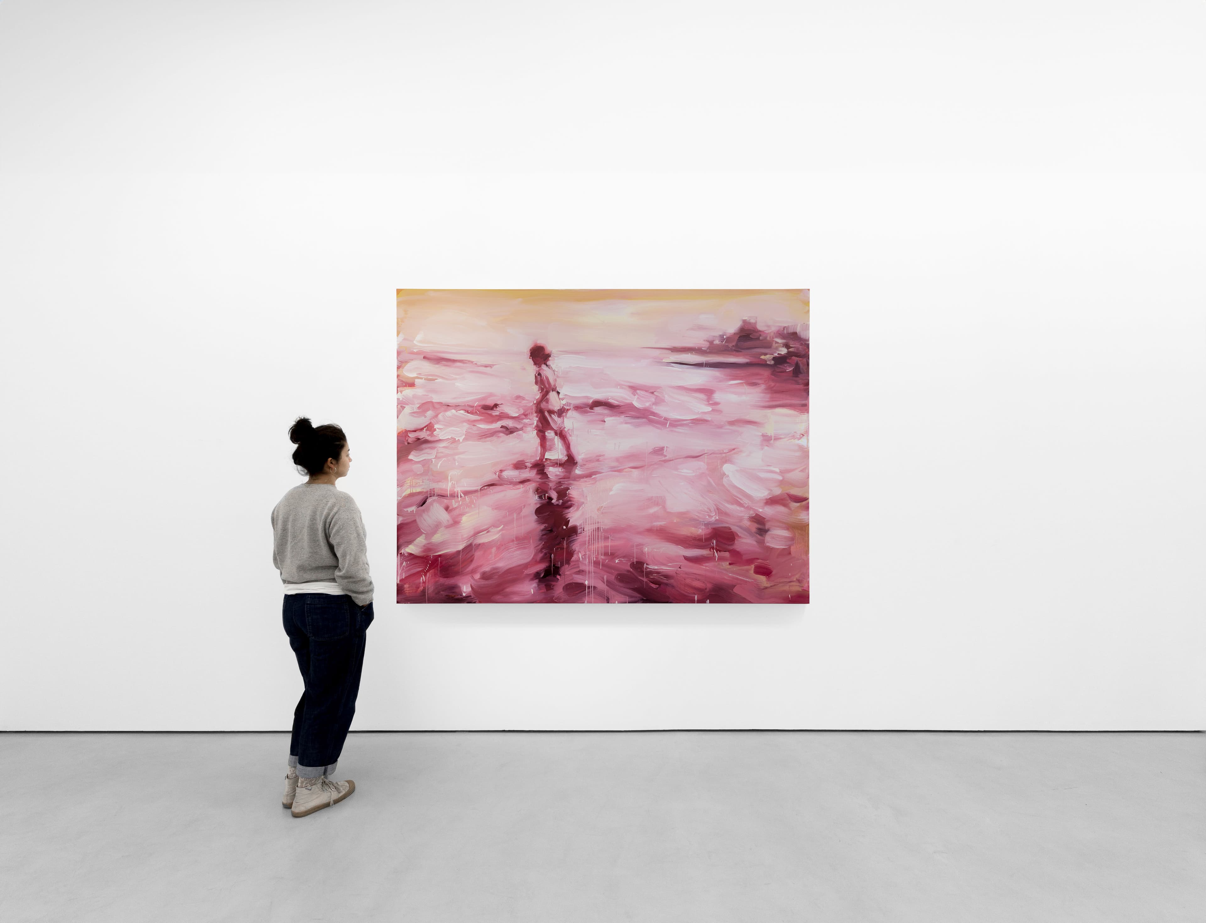 installation shot of a figue standing in front of a loosely painted landscape painting by Laura Lancaster - a woman walks to the left across wet sand towards the sea, the coastline is visible in the top right hand corner. The scene is bathed in vibrant pinks and yellowshed  