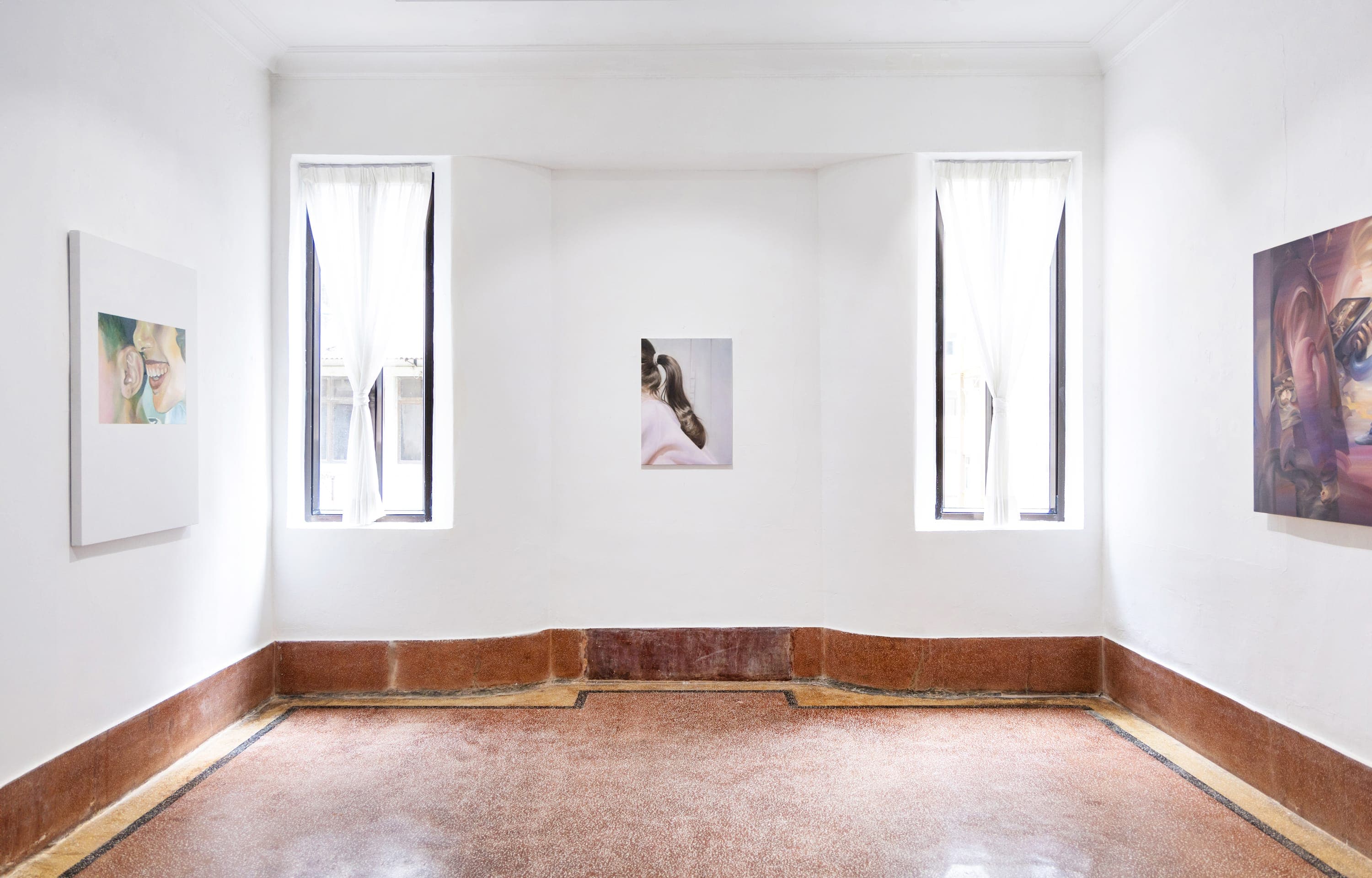 A white walled gallery with three artworks hung on the walls and two slim windows 