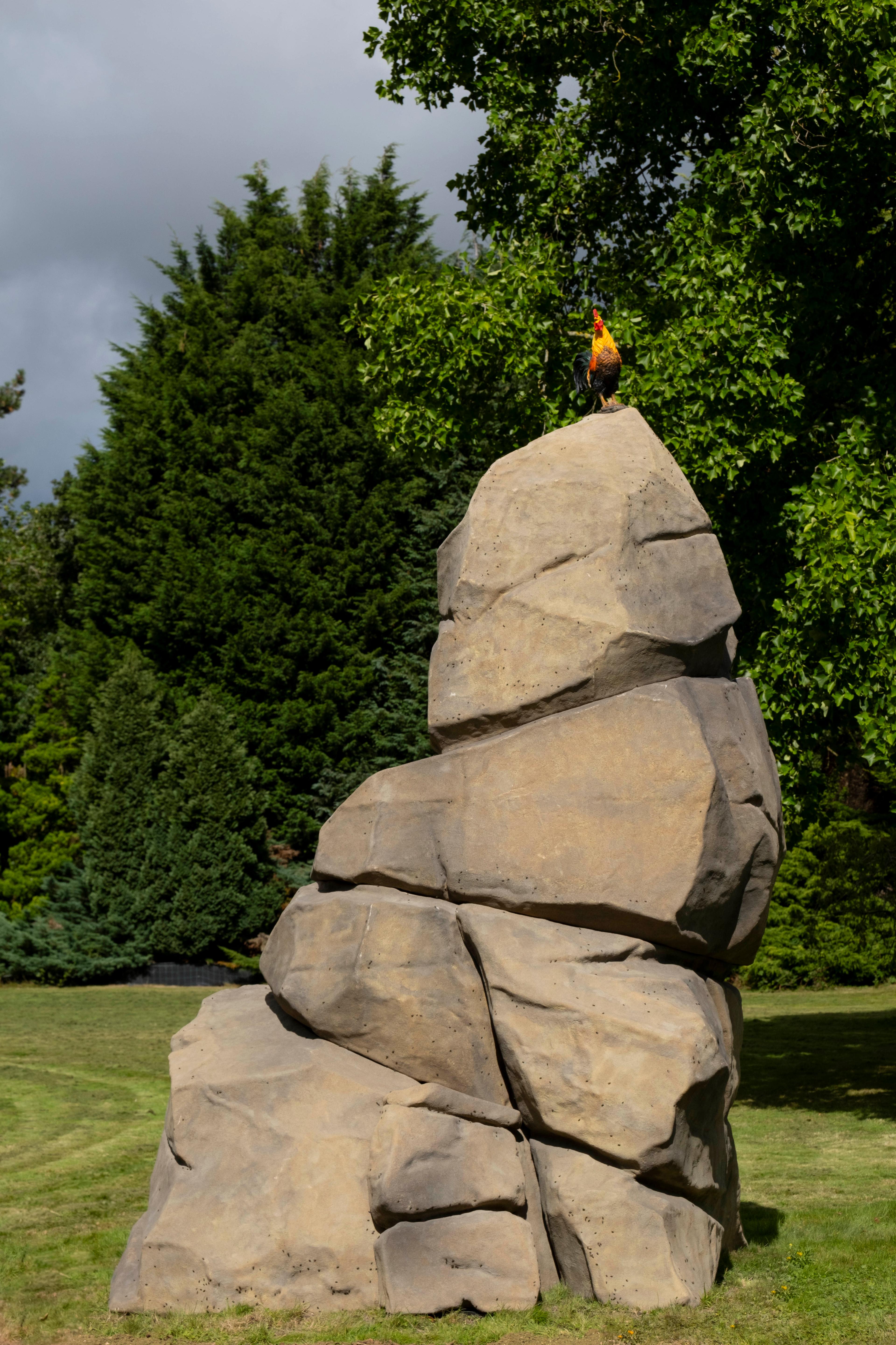 A large sculpture by Simeon Barclay of a large rock with a cockerel sitting on top placed in a grassy field