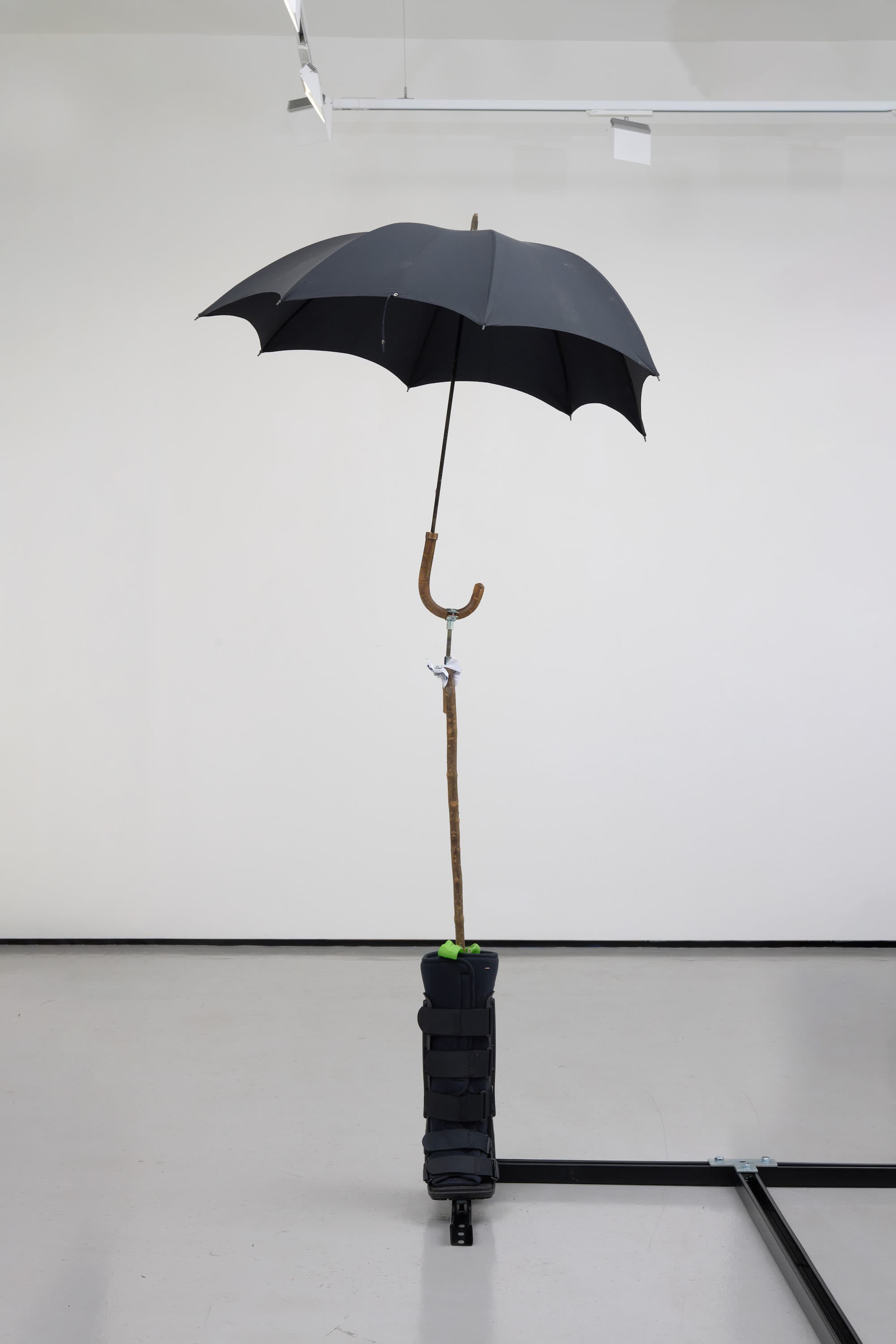 an image of Simeon Barclay's sculpture 'Warrior Falling on Their Feet', 2023 consiting of a Walking stick, umbrella, concrete, betting slip, foot fracture brace walker, bag for life