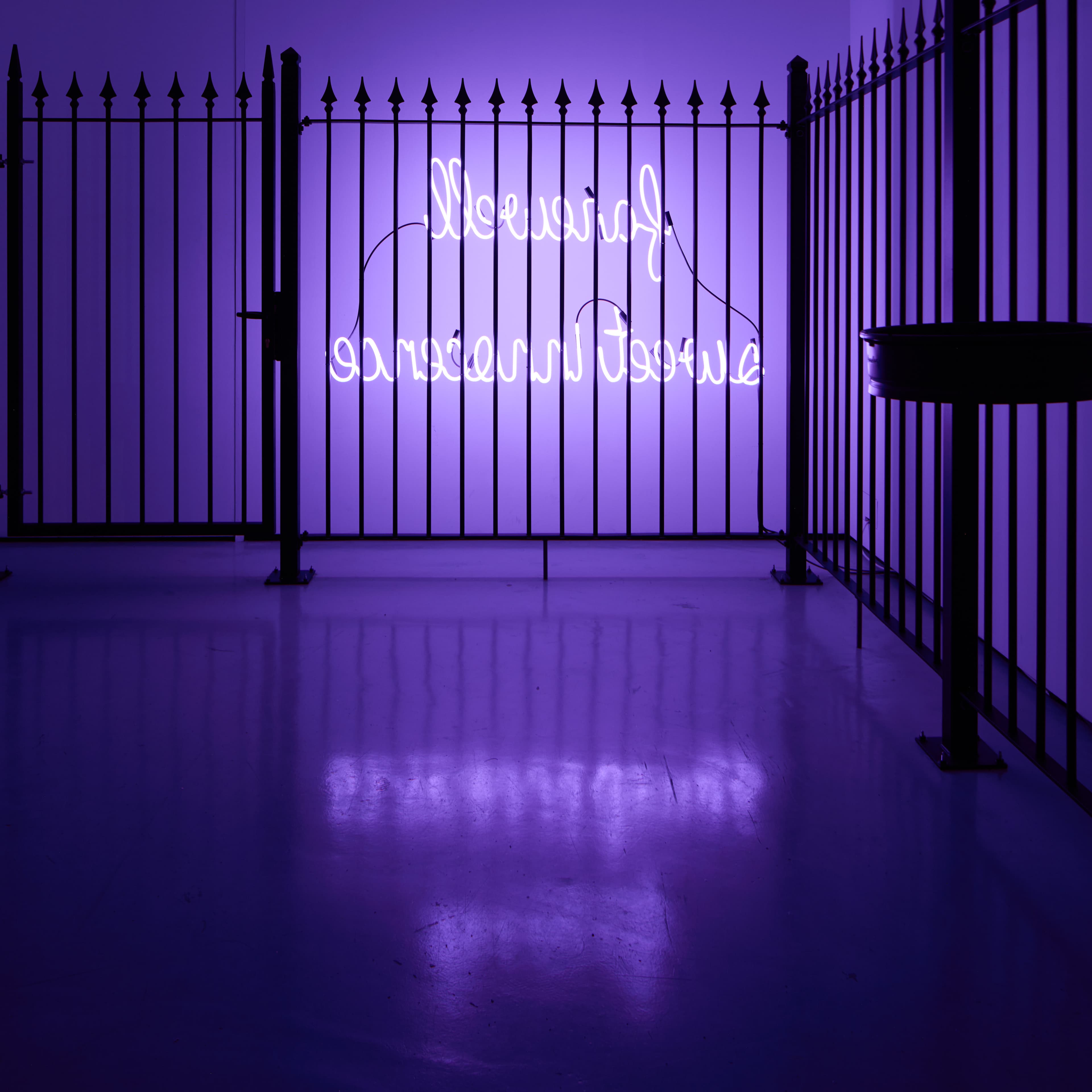 Photograph of ɘɔnɘɔonni Ɉɘɘwƨ llɘwɘяɒʇ, 2023 by Simeon Barclay, consiting of a Metal fence, neon, plastic, metal bin holders, metal fixings. The black metal railings are lit by the purple glow of the neon text. 