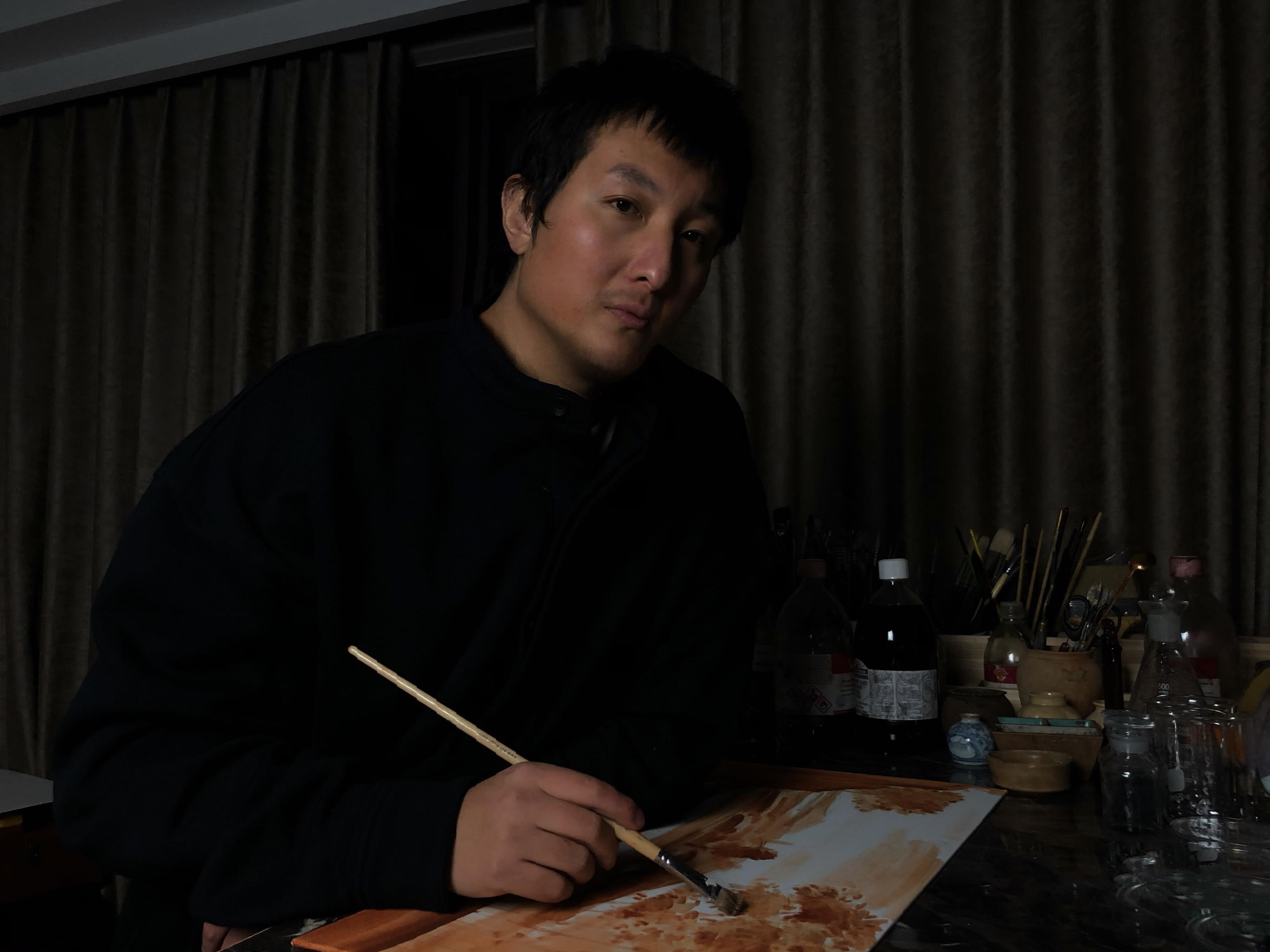 A portrait of Pei Wang sat in a dark room holding a paintbrush