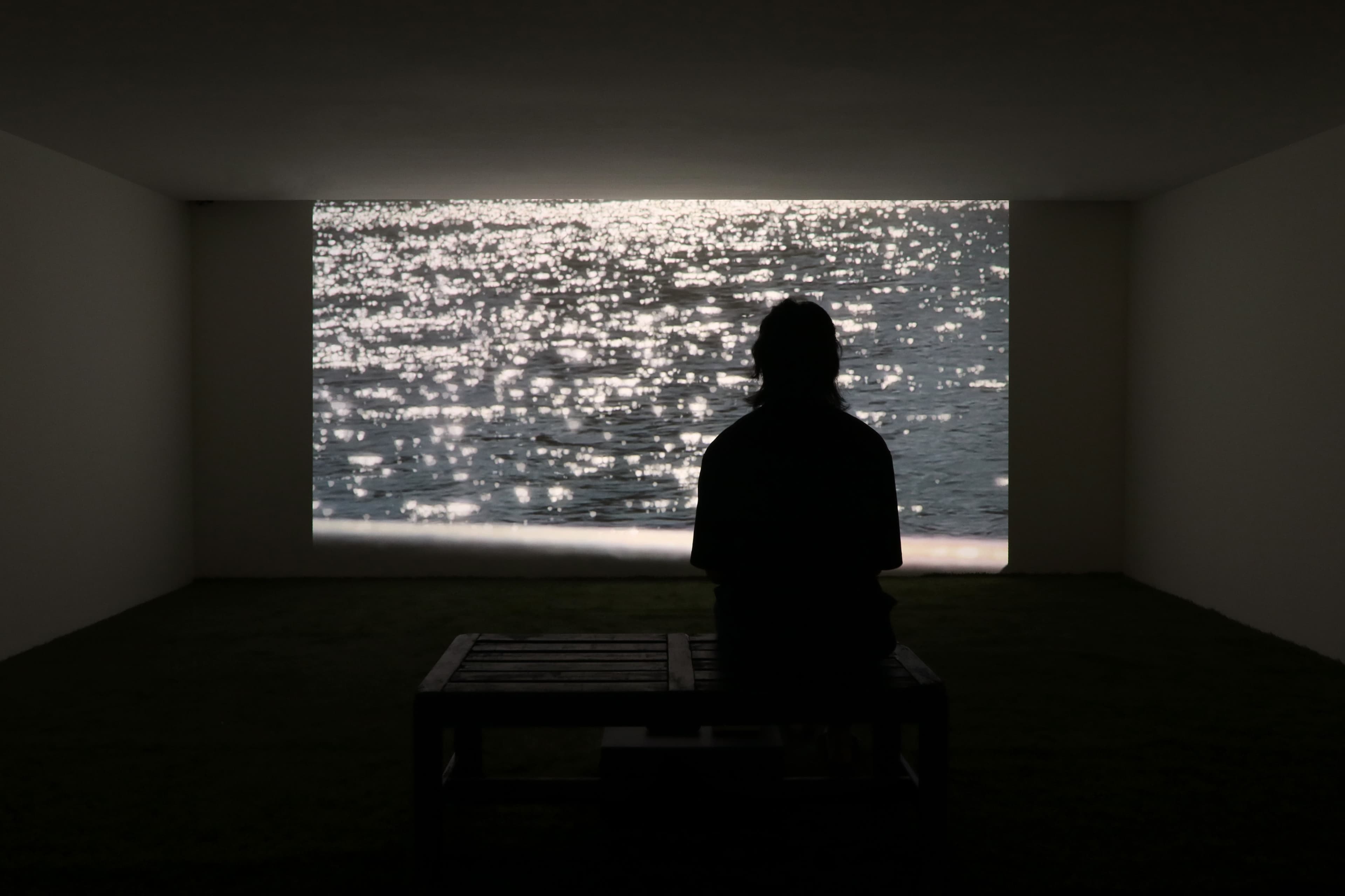 Installation view of a dark room with a figure silhouetted against a large projection of a film by James Prapaithong