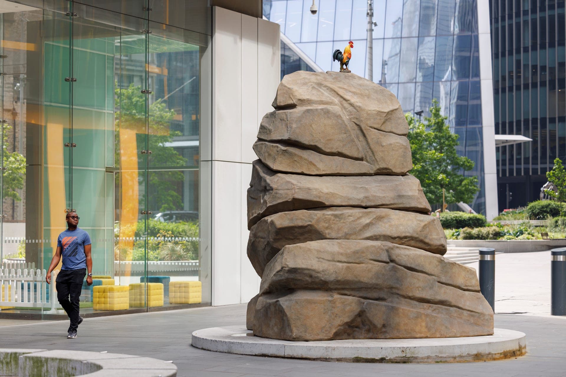 A man walking past a large sculpture by Simeon Barclay of a boulder with a cockerel stood on top