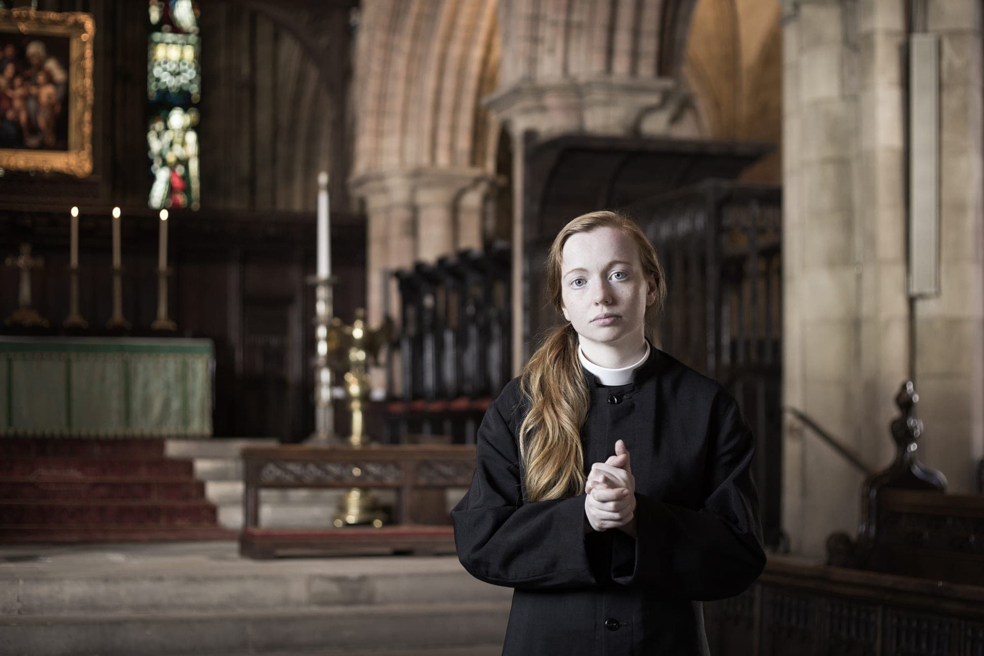 An image from Matt Stokes' film 'This Liberty' - a female priest looks at us from within a cathedral setting. She has her hands clasped.