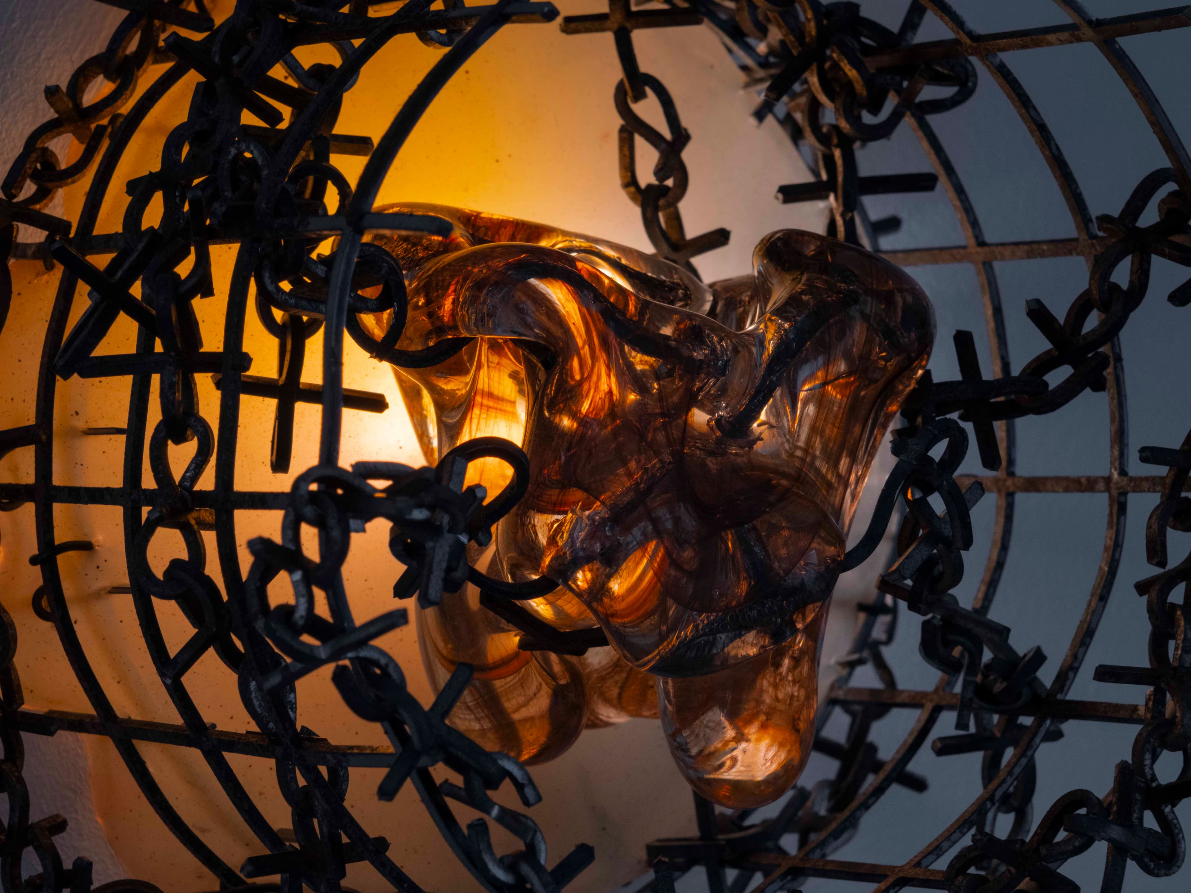 Wall sculpture by Hazel Brill. Circular metal frame intertwined with barbed metal chain which hangs below and an orange centre glow of a resin globby drip.