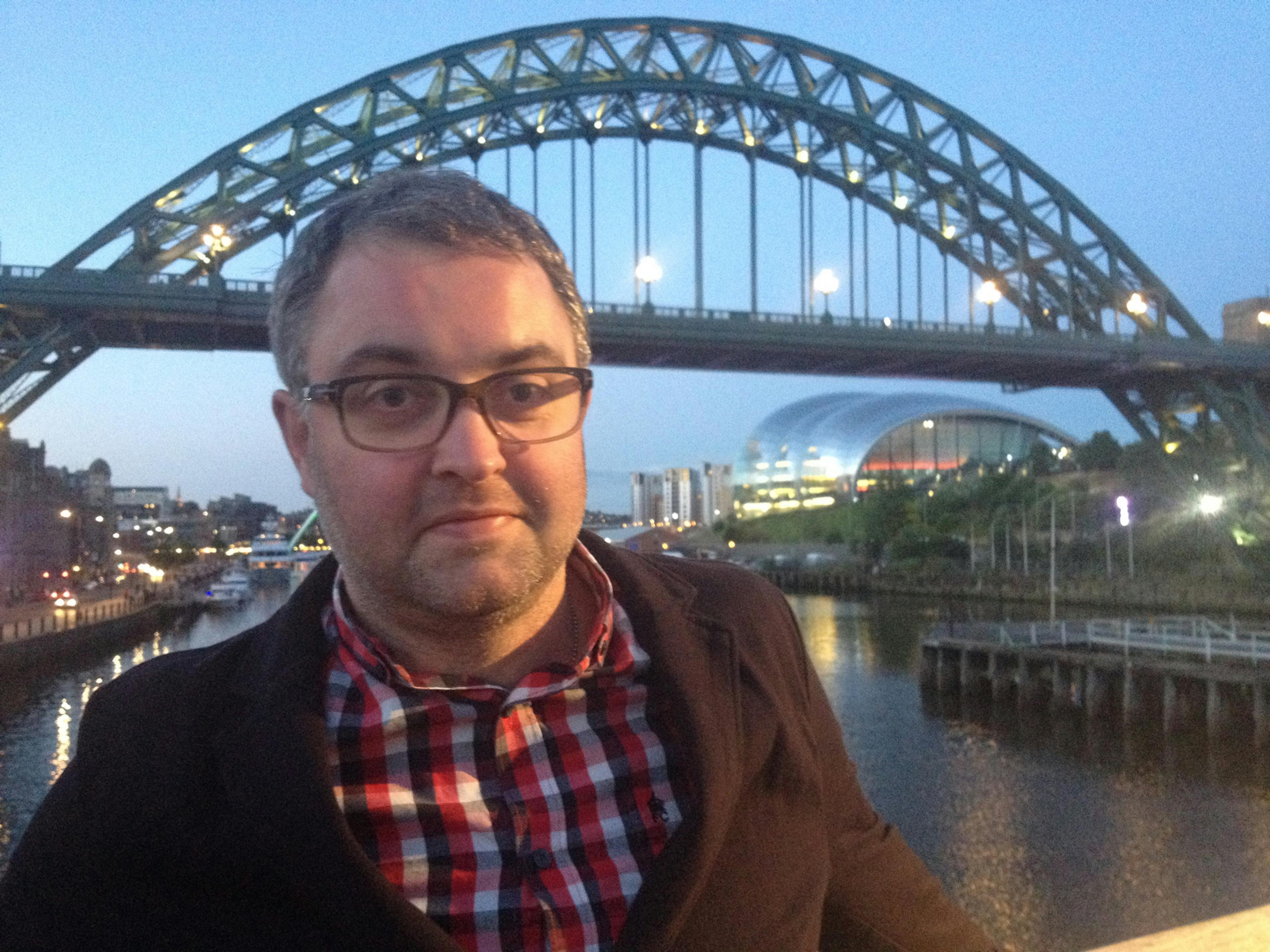 A photograph of Paul Moss stood in front of the Tyne Bridge and Gateshead quays. He is wearing a red and white checked shirt and glasses.