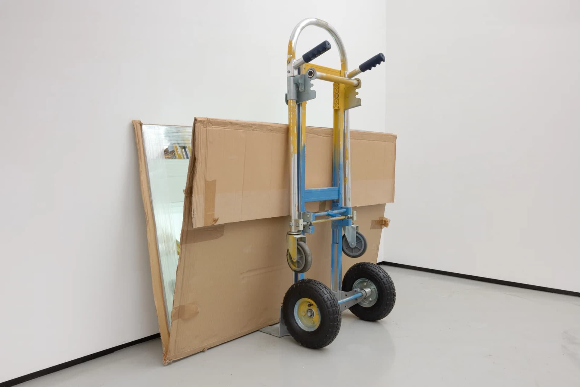 A sculpture by Rafael Pérez Evans consisting of a yellow and blue parcel truck resting against the wall with mirrors packaged in cardboard propped open at 15 degrees with yams, making an wheel of yams