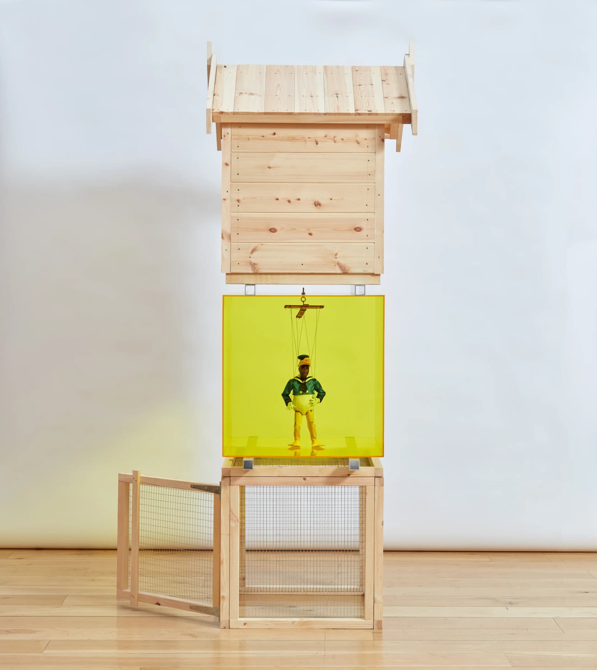 Iceberg is a sculpture by Simeon Barclay comprised of a stack of a hutch, a yellow perspex cube with a wooden puppet of the artist in Elton John's 'Donald Duck' costume and a dog kennel