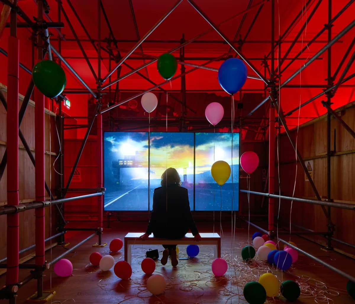 A photo of a woman watching a cinema screen in a red light room with scaffolding and balloons.