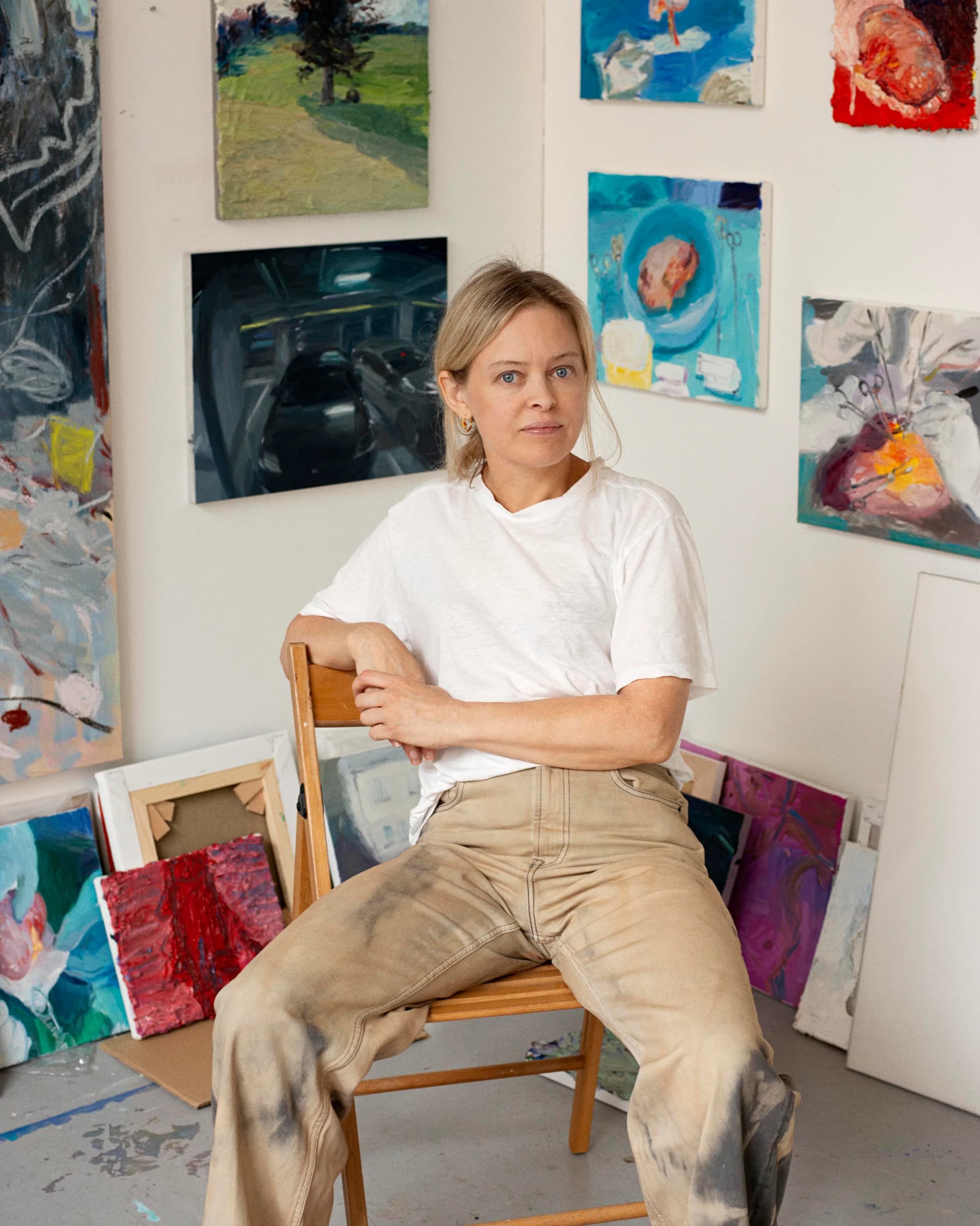 A portrait of Celia Hempton sat on a chair in her studio wearing a white t-shirt and beige trousers