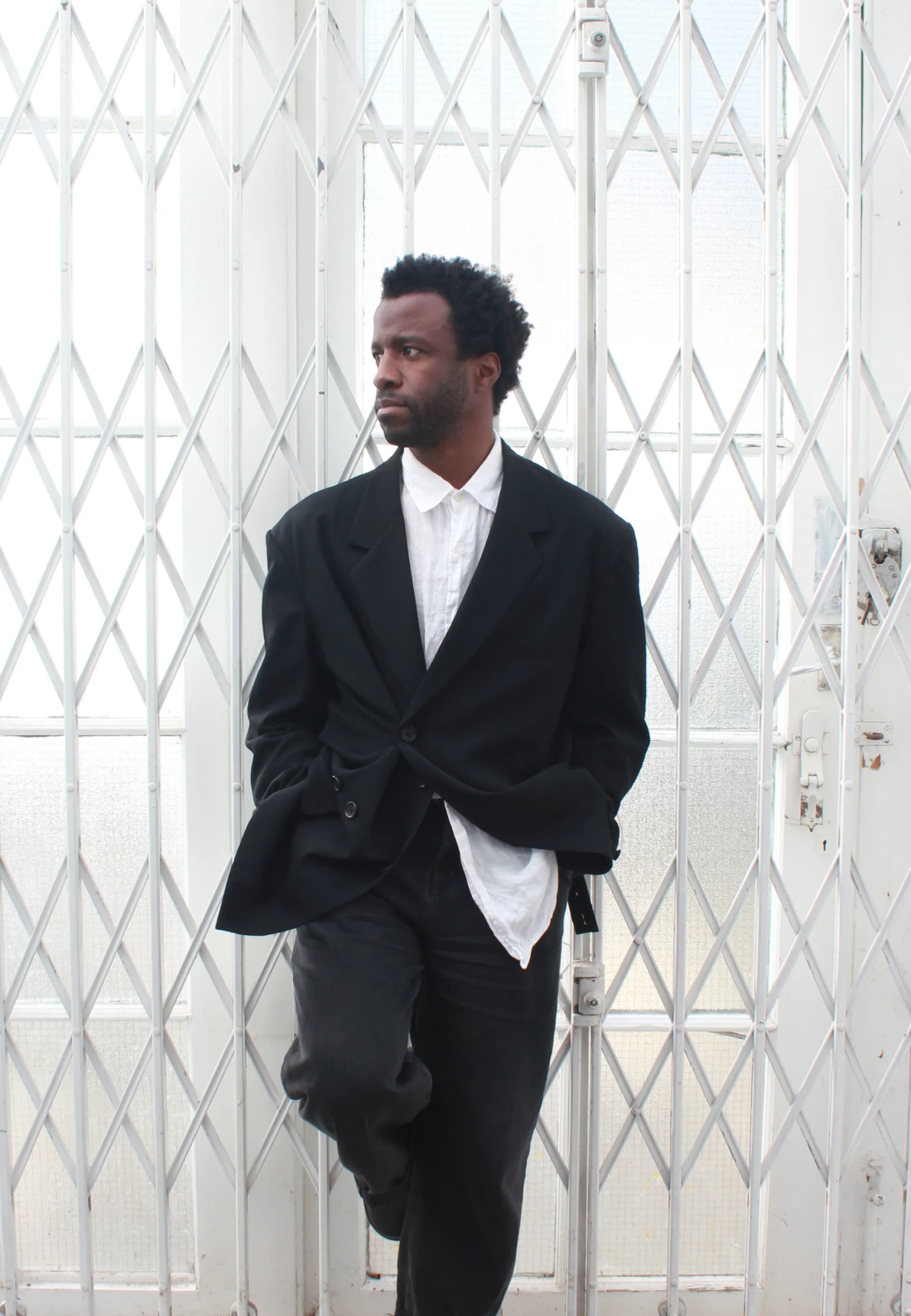 Portraits of artist Simeon Barclay wearing a black suit with white shirt in a minimal semi-industrial setting