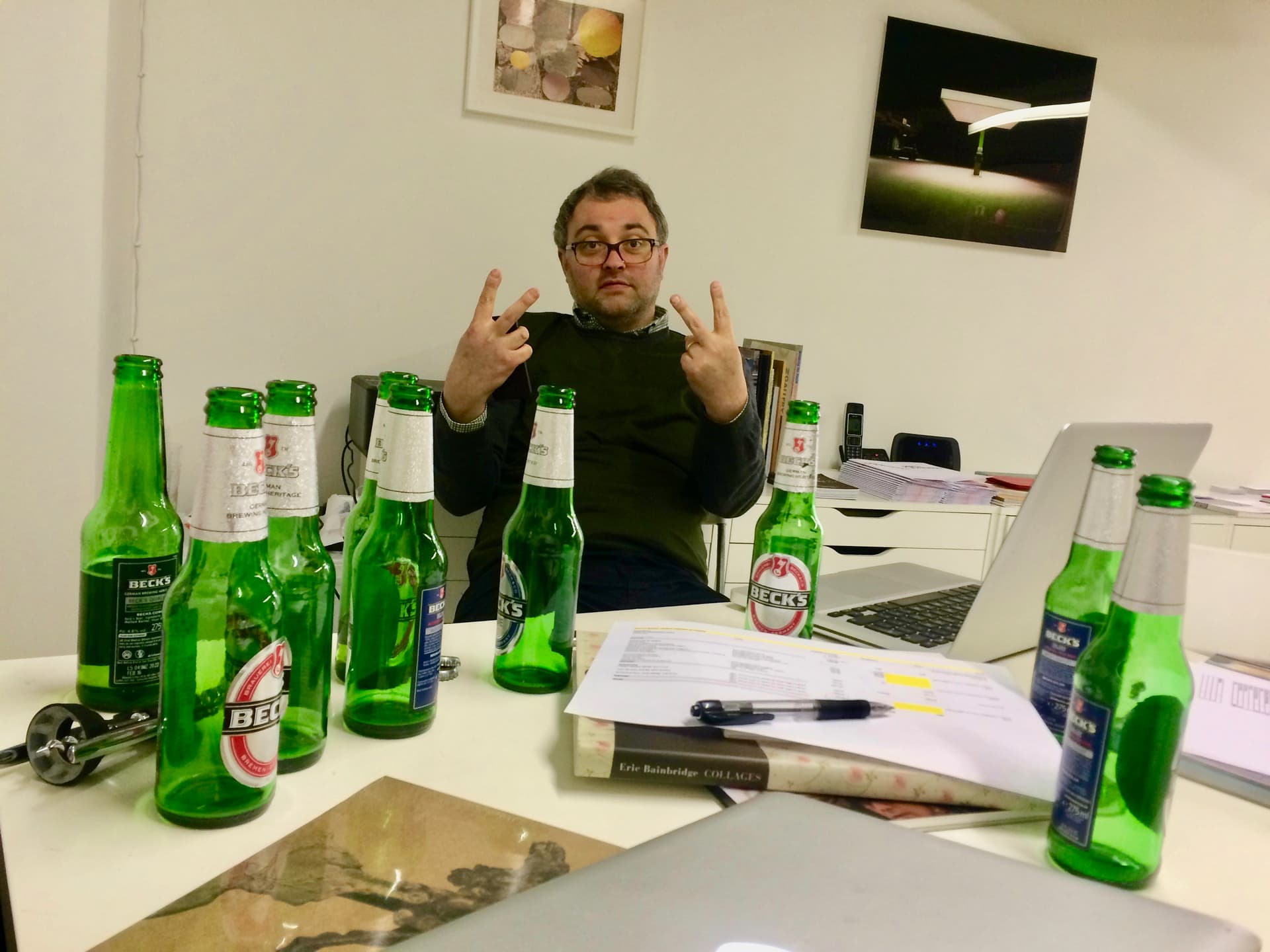 Paul Moss photographed in the gallery office. He is staring directly at the camera and is giving the double V sign. In front of him are several empty bottles of beer.behind many